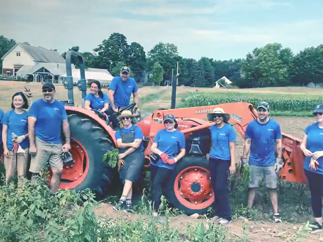 A group of people standing next to or on a large farm tractor in a field of growing crops. A home behind them. They're all wearing a blue shirt with "Alkermes" on it.