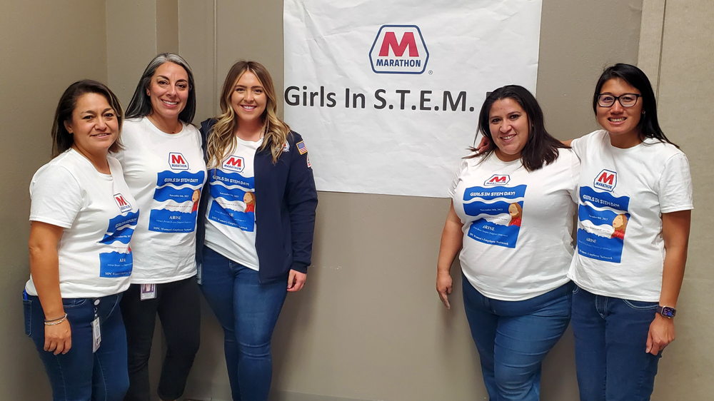A group of five women with arms around each other in front of a banner "Girls in S.T.E.M." and marathon logo. Each wears a Marathon t-shirt.
