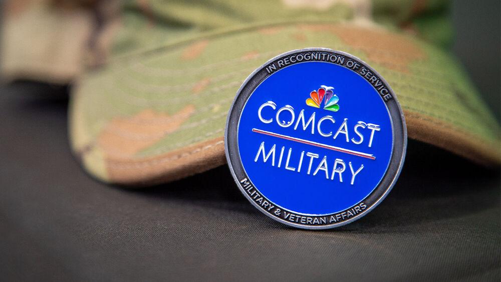 Pin with Comcast Military on it. A camouflage hat behind it. 
