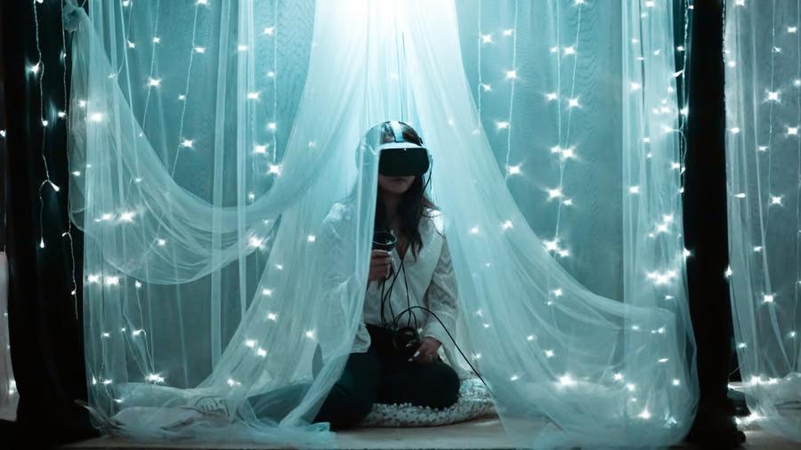 A person using a VR headset and controllers, sheer drapes and string lights around them.