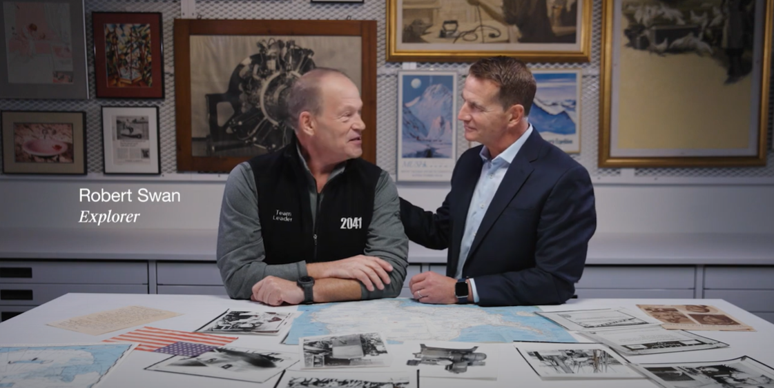 Robert Swan and Kohler representative standing in front of a table with scattered photos from Robert's expeditions. Old pictures of the arctic and explorers hanging on the wall behind them.