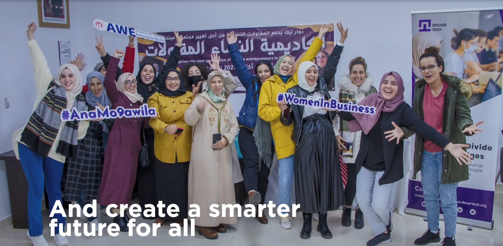 A group of women, smiling, with arms raised, most wearing head-coverings. A sign in a foreign language behind them, and some holding cutout hashtag references.