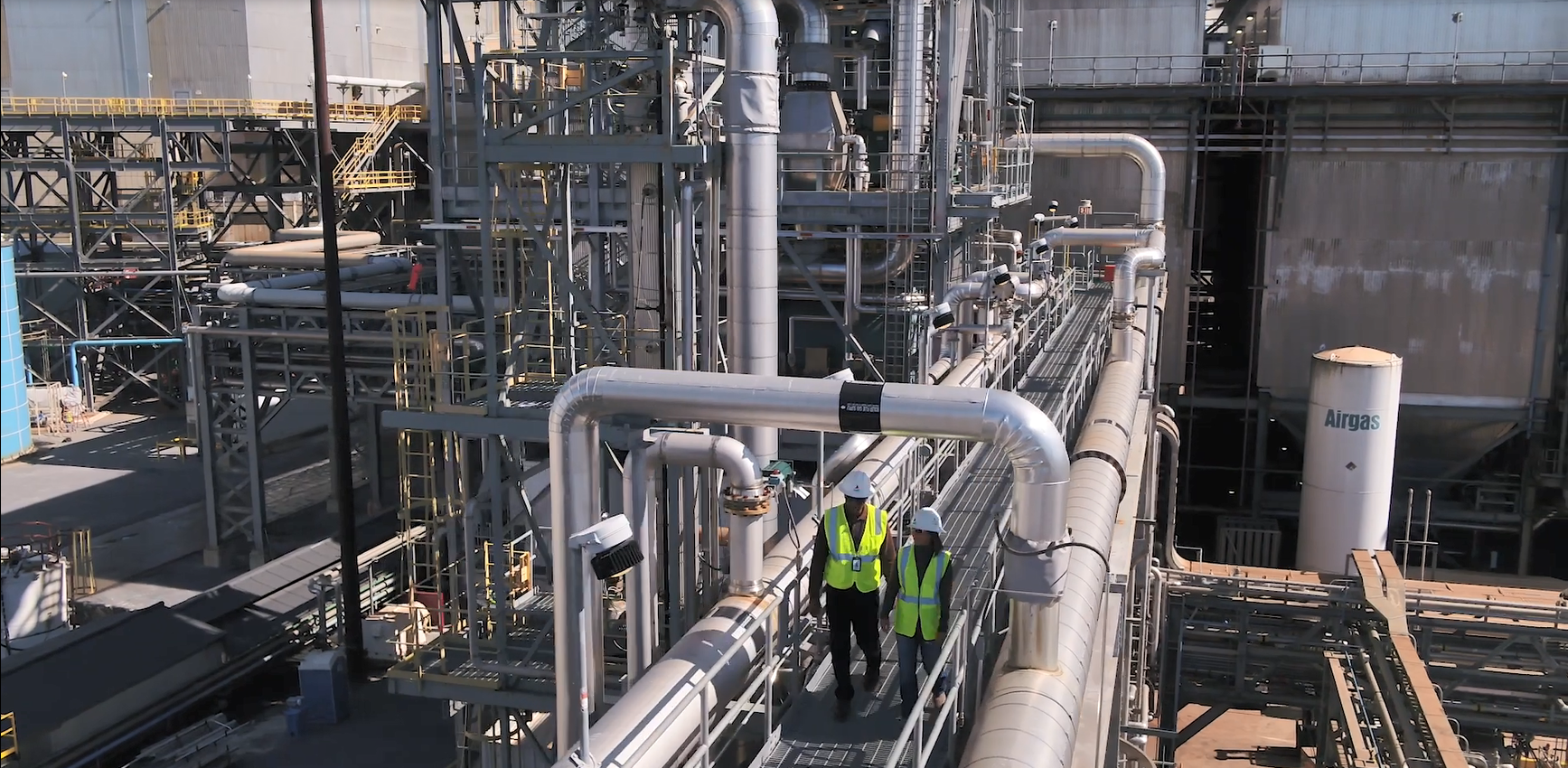 Two people wearing hard hats and high-vis vests walking on a long elevated pathway. Industrial piping and building behind them.