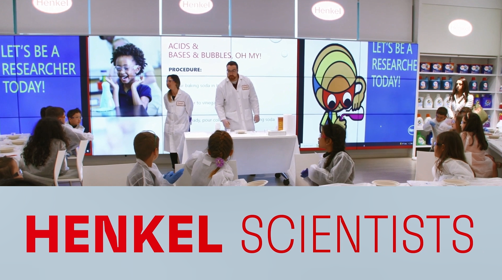 Two scientists in lab coats in front of a room of seated children. "Henkel Scientists" at the bottom.