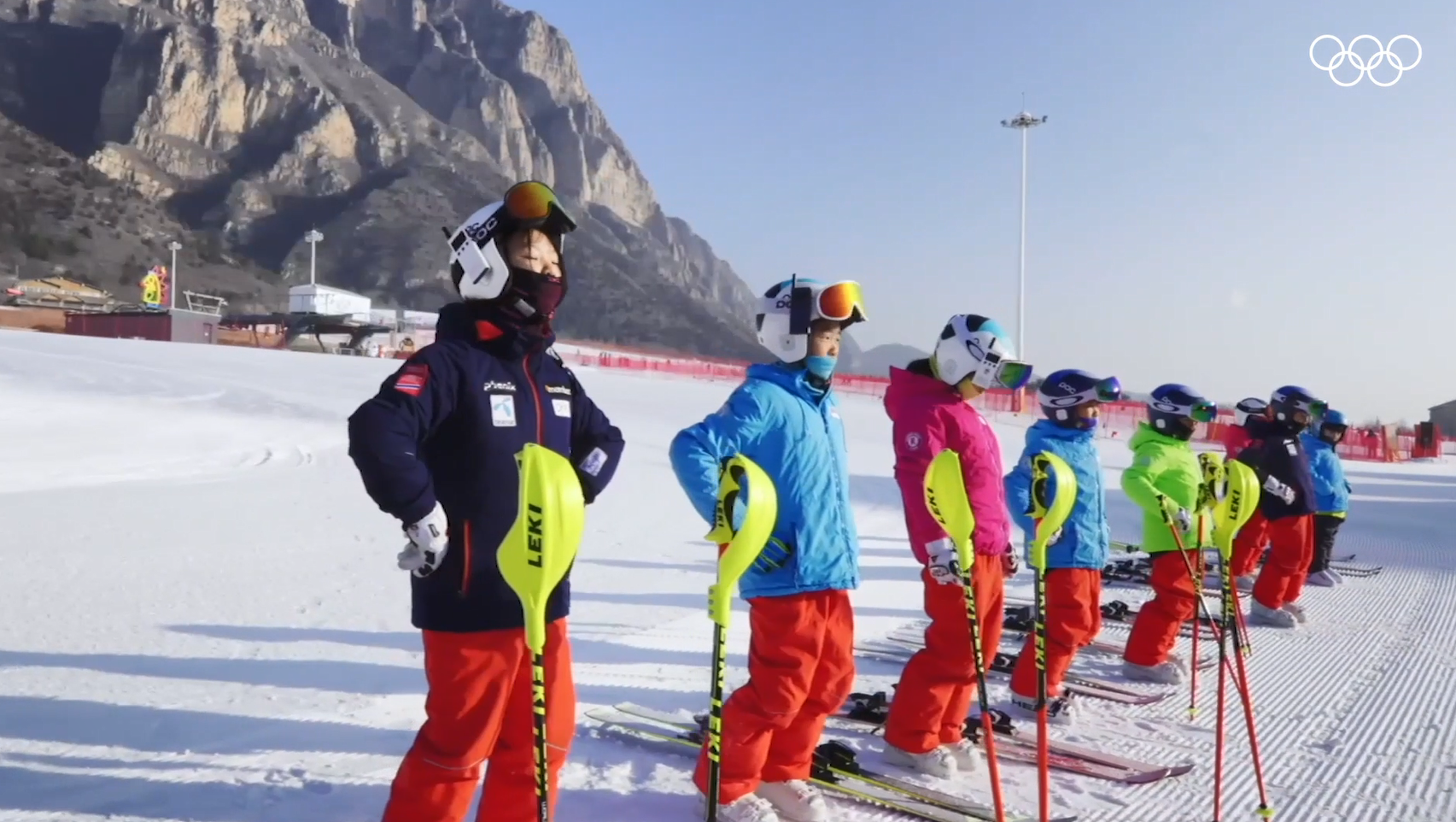 Children in a line do stretches on skis on a snowy hill.