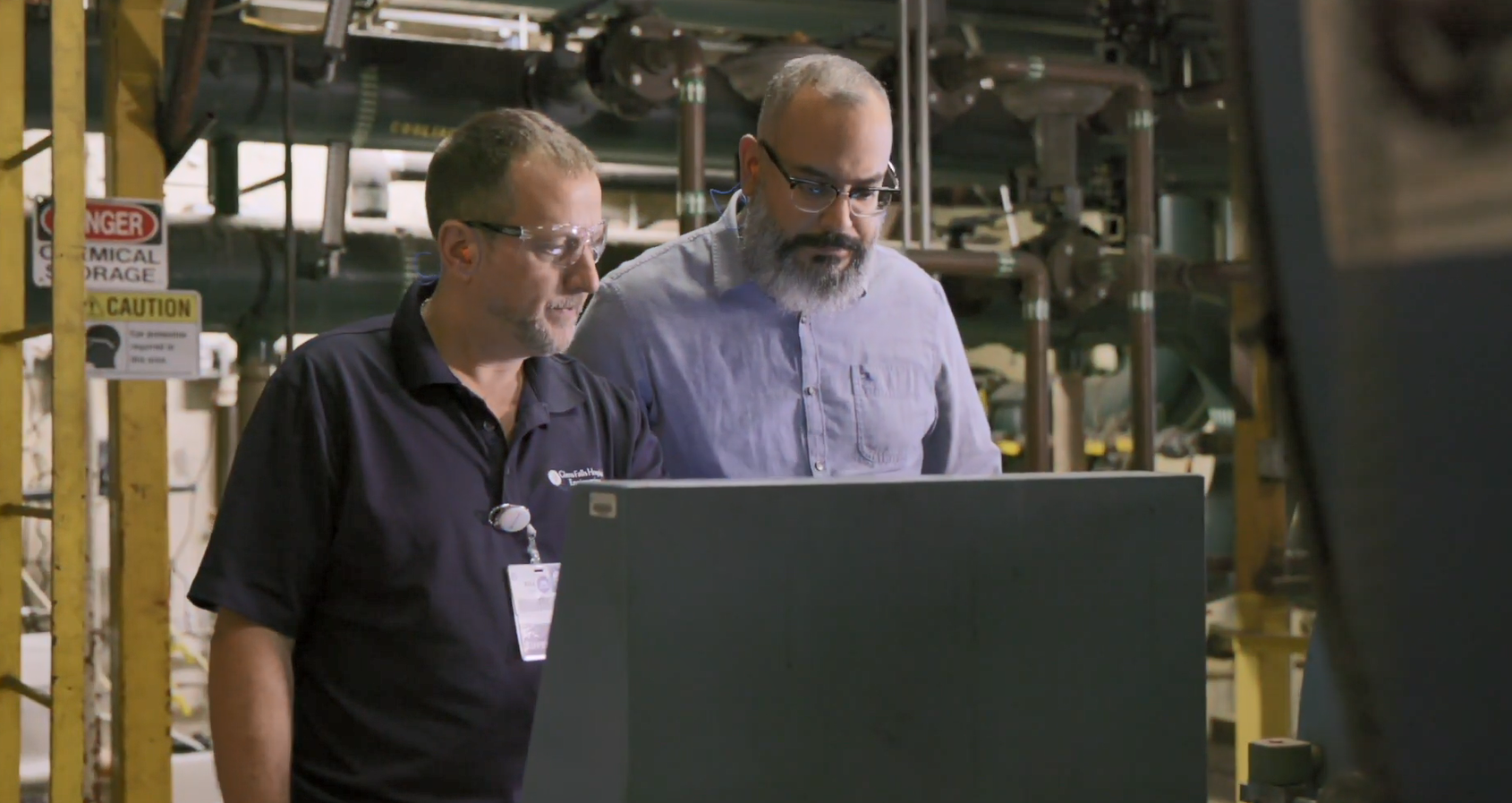 Two people looking at a screen in an room with may pipes and wires.