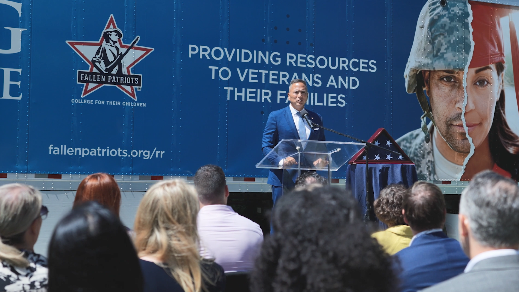 A person speaking at a podium. An american flag in a triangle box to the right and a truck behind covered with "Fallen Patriots" logos and pictures of veterans and service members.