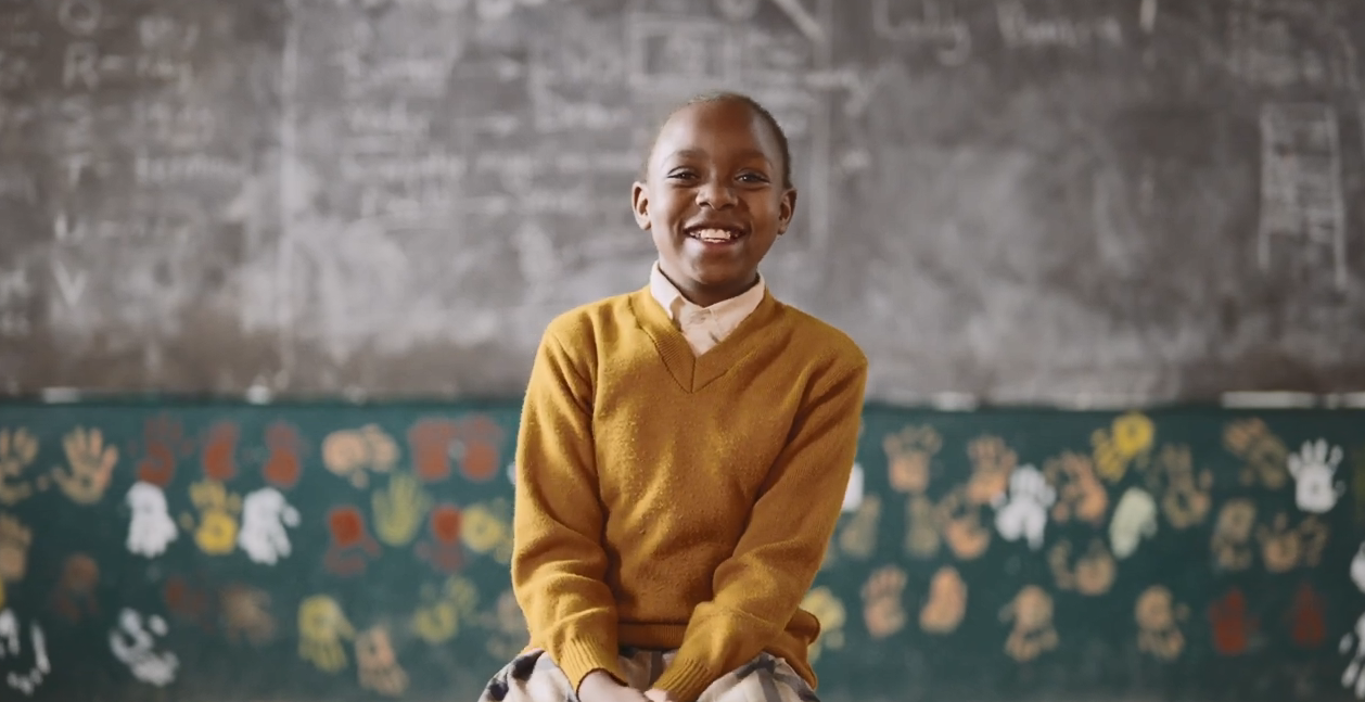 A student smiling, seated at the front of a classroom.