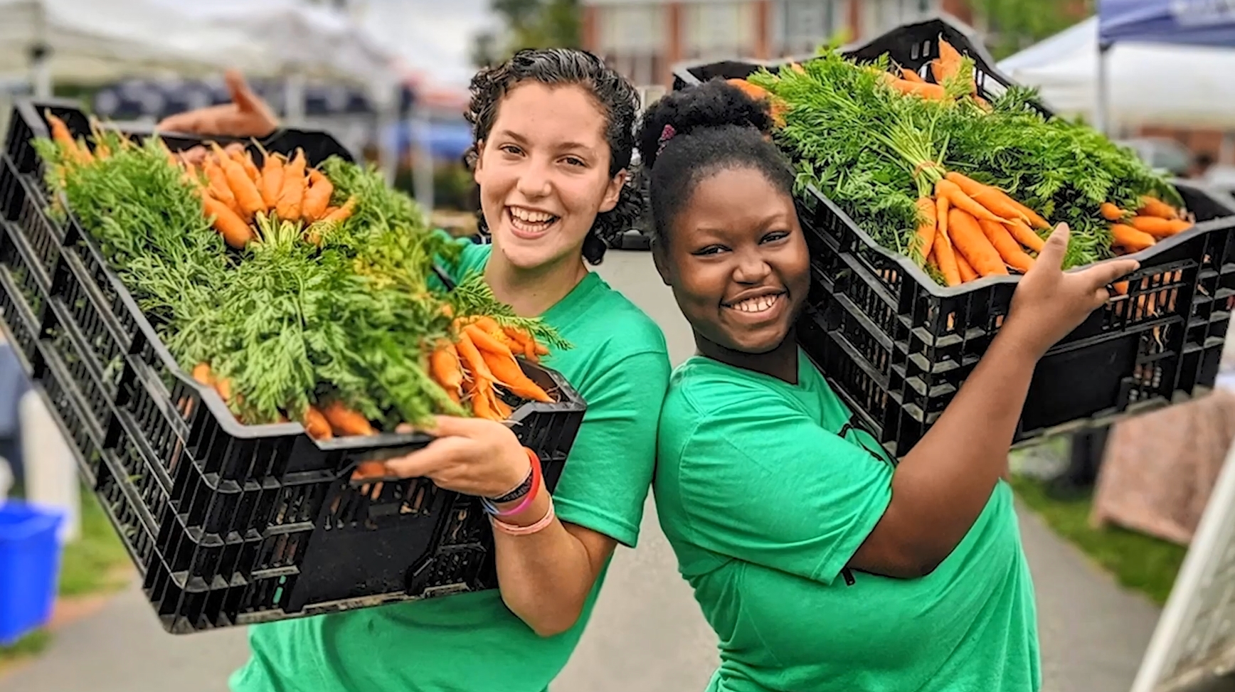 Two smiling people holding up large bins of fresh carrots.