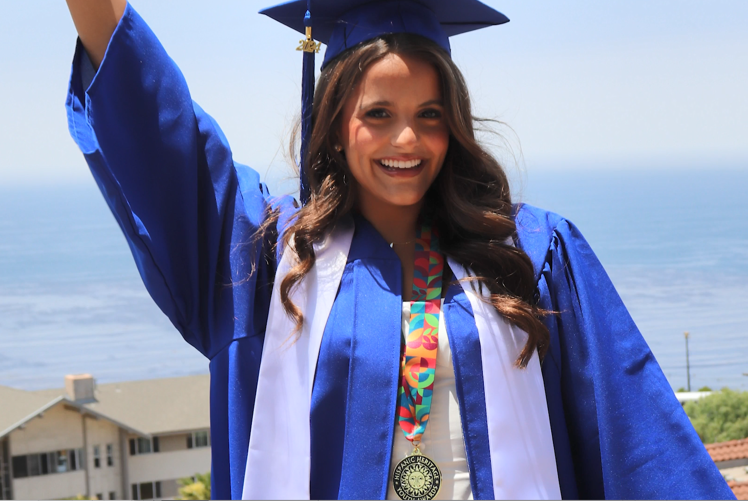 A smiling person in graduation cap and gown, one arm raised high. A scenic expanse of water behind them.
