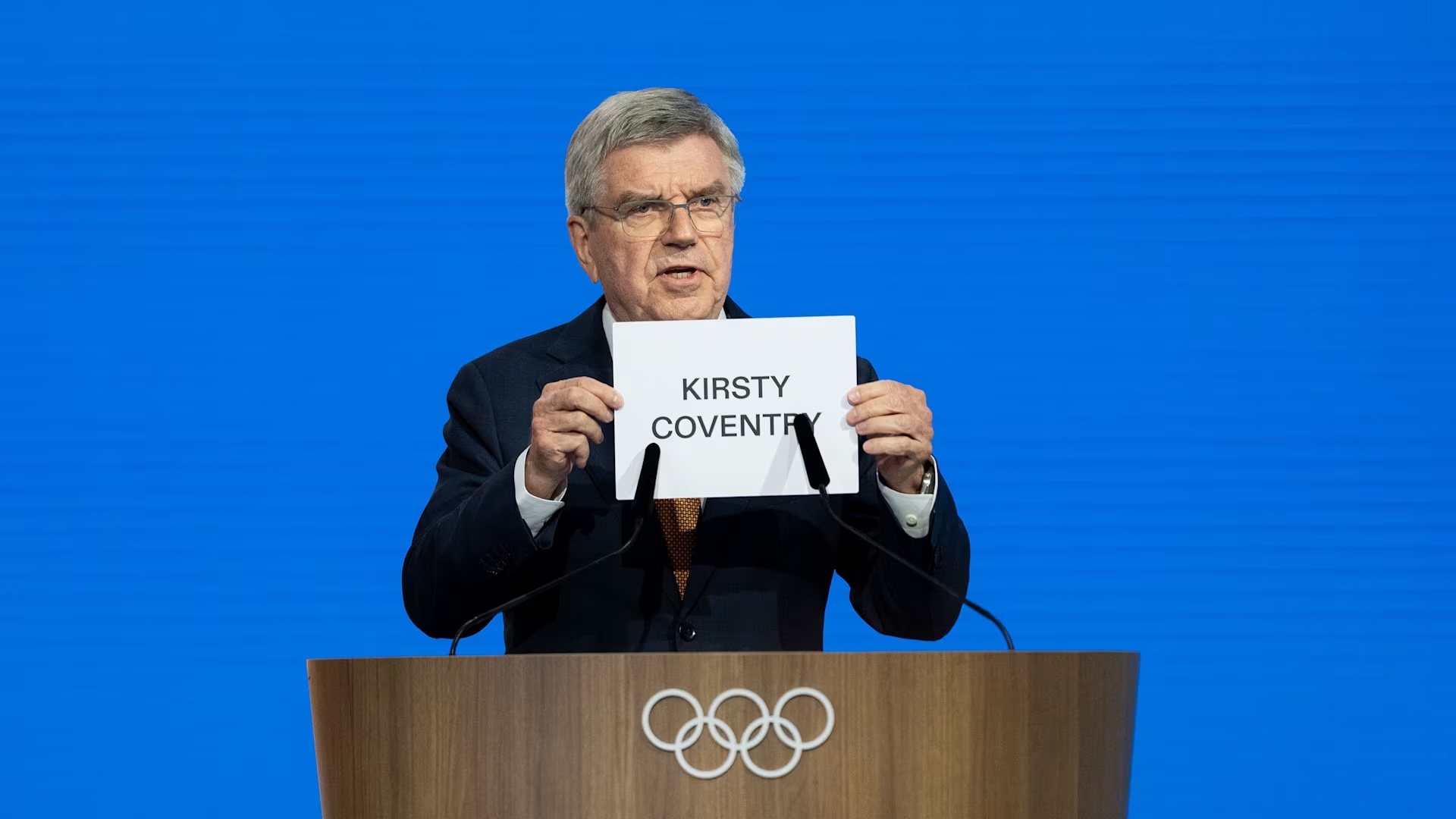 Man at lectern holding up s sign reading, "Kirsty Coventry"