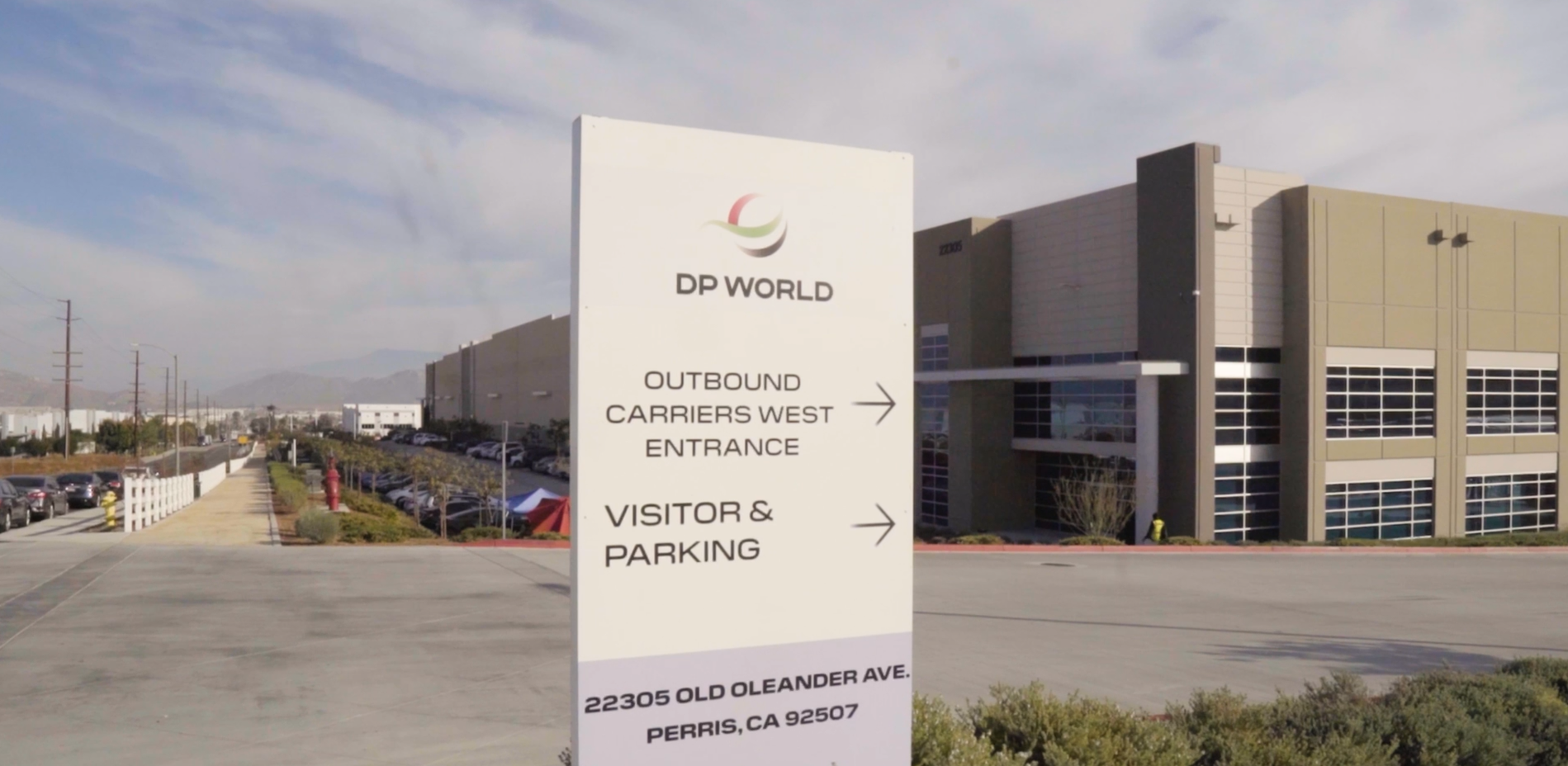A DP World logistics facility in Perris, California, featuring a sign at the entrance directing outbound carriers and visitors to parking, with the warehouse and loading areas visible in the background under a clear sky.
