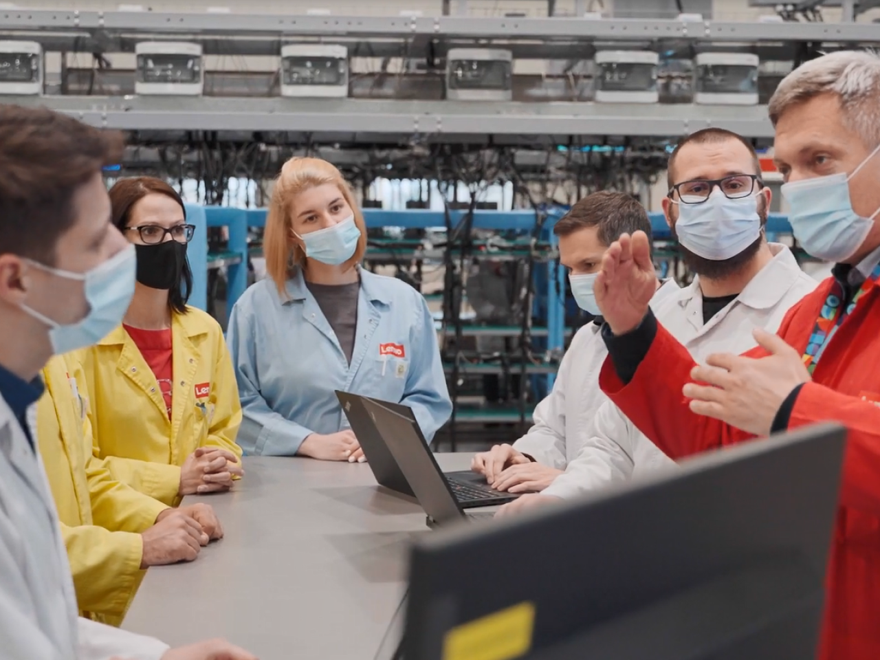 small group of people gathered around a table, all wearing protective masks and lab coats of different colors. Laptops open on the table. Factory machinery behind them.