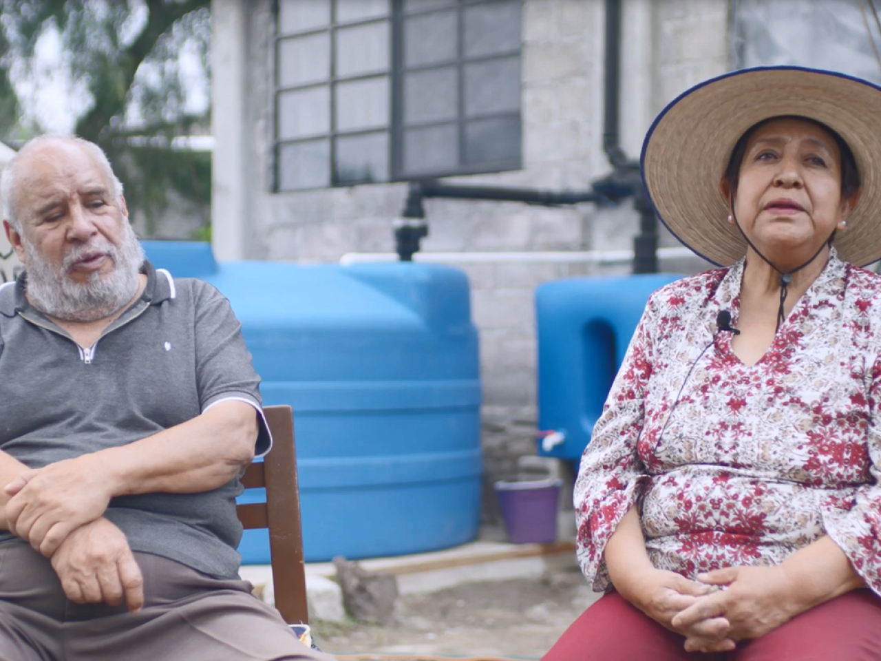 Gloria and Sergio sitting in chairs outside, in front of large water collection containers.