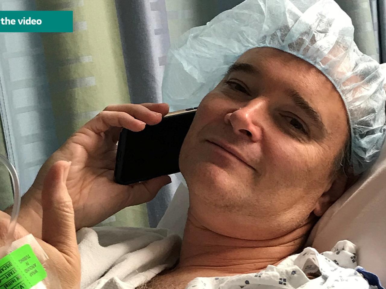Chet Kitchen on a cell phone, wearing a hospital gown and hair covering.