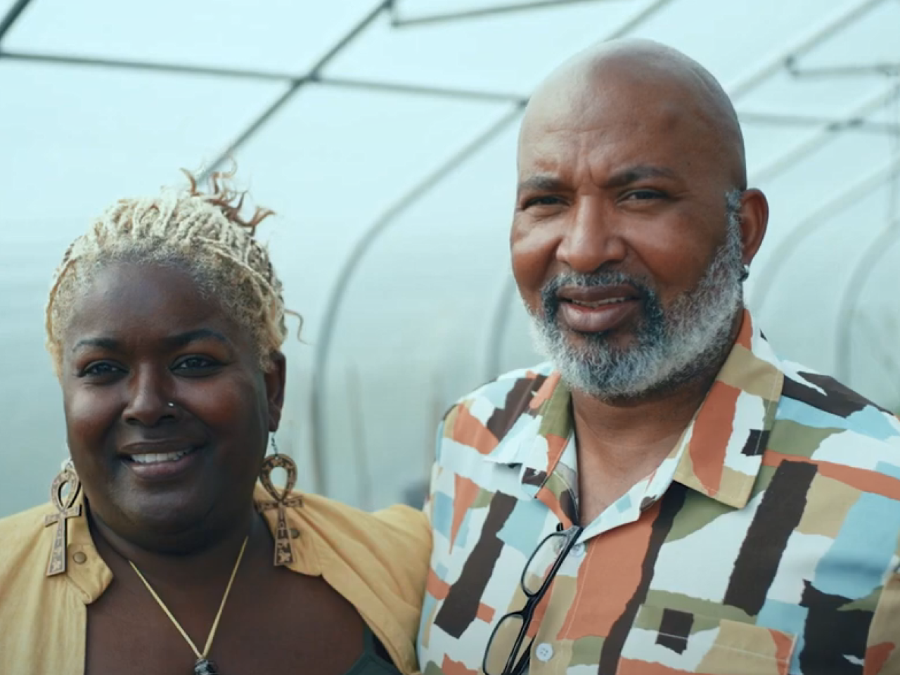 Two people standing in a greenhouse, an arm around each other.