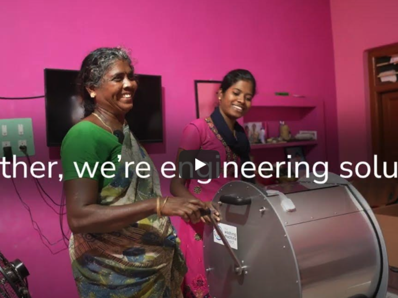 Two people with a washing machine in front of a bright pink wall