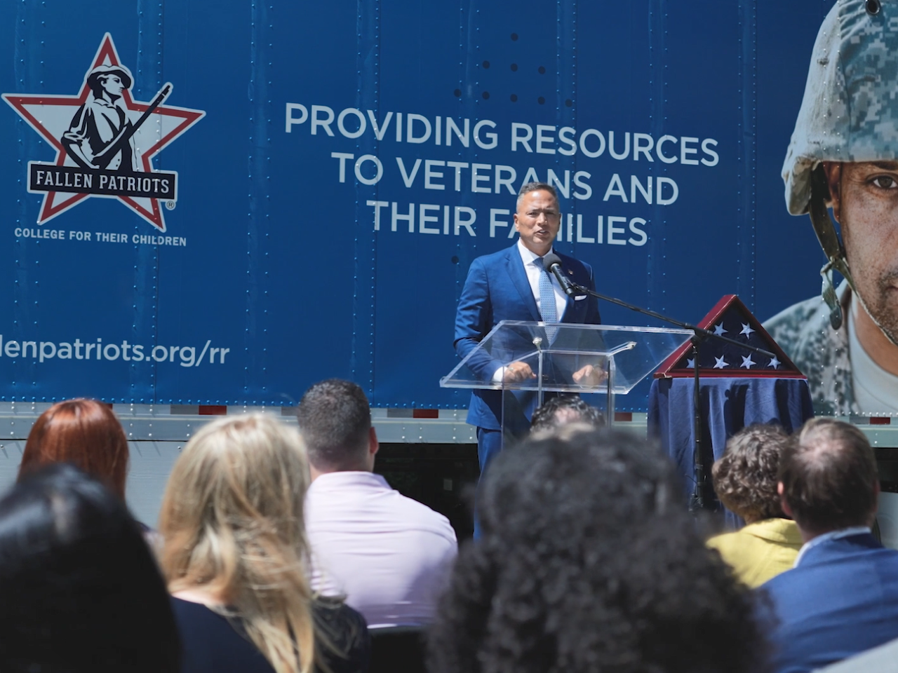 A person speaking at a podium. An american flag in a triangle box to the right and a truck behind covered with "Fallen Patriots" logos and pictures of veterans and service members.