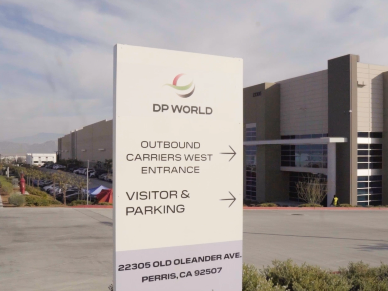 A DP World logistics facility in Perris, California, featuring a sign at the entrance directing outbound carriers and visitors to parking, with the warehouse and loading areas visible in the background under a clear sky.
