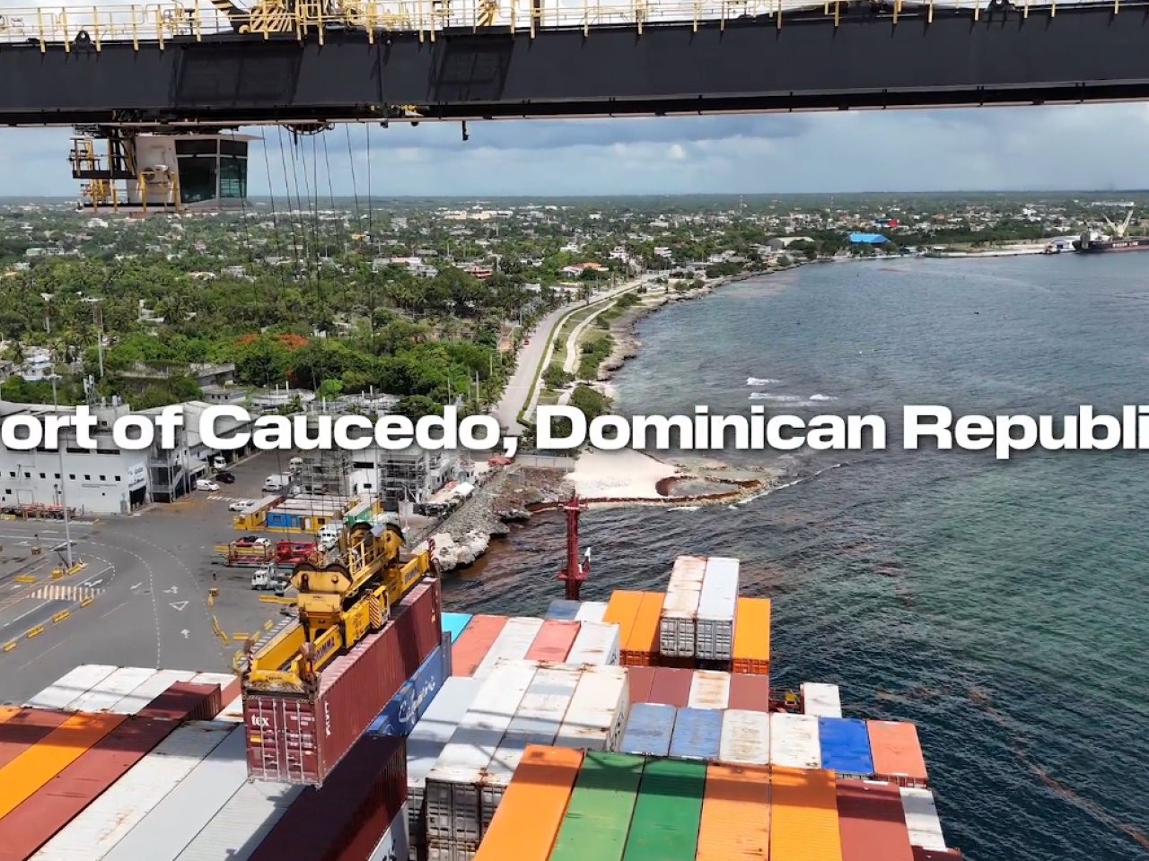 Aerial view of the Port of Caucedo in the Dominican Republic, showing container operations along the coastline with cranes lifting cargo, stacked shipping containers, and the Caribbean Sea in the background.
