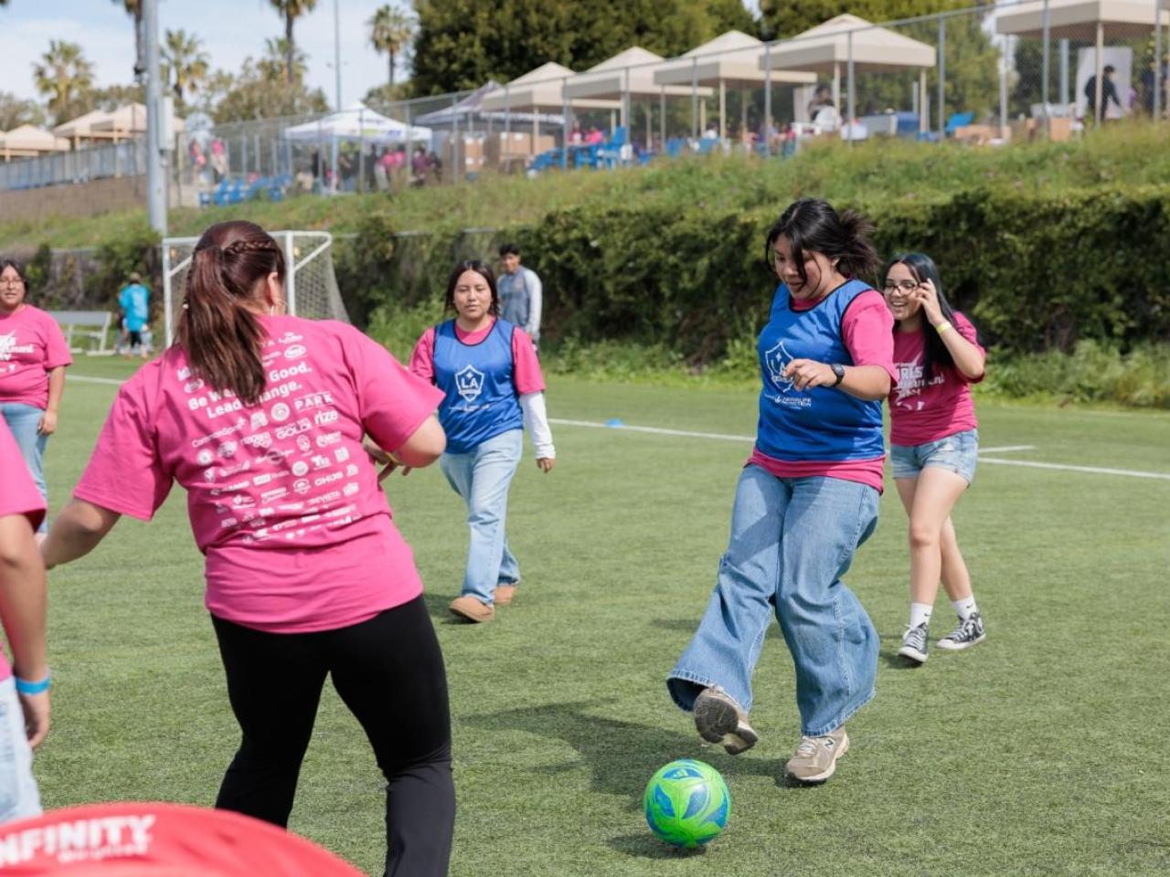 Girls wearing pink Girls Empowerment Day shirts play soccer during a youth sports clinic on an outdoor field.