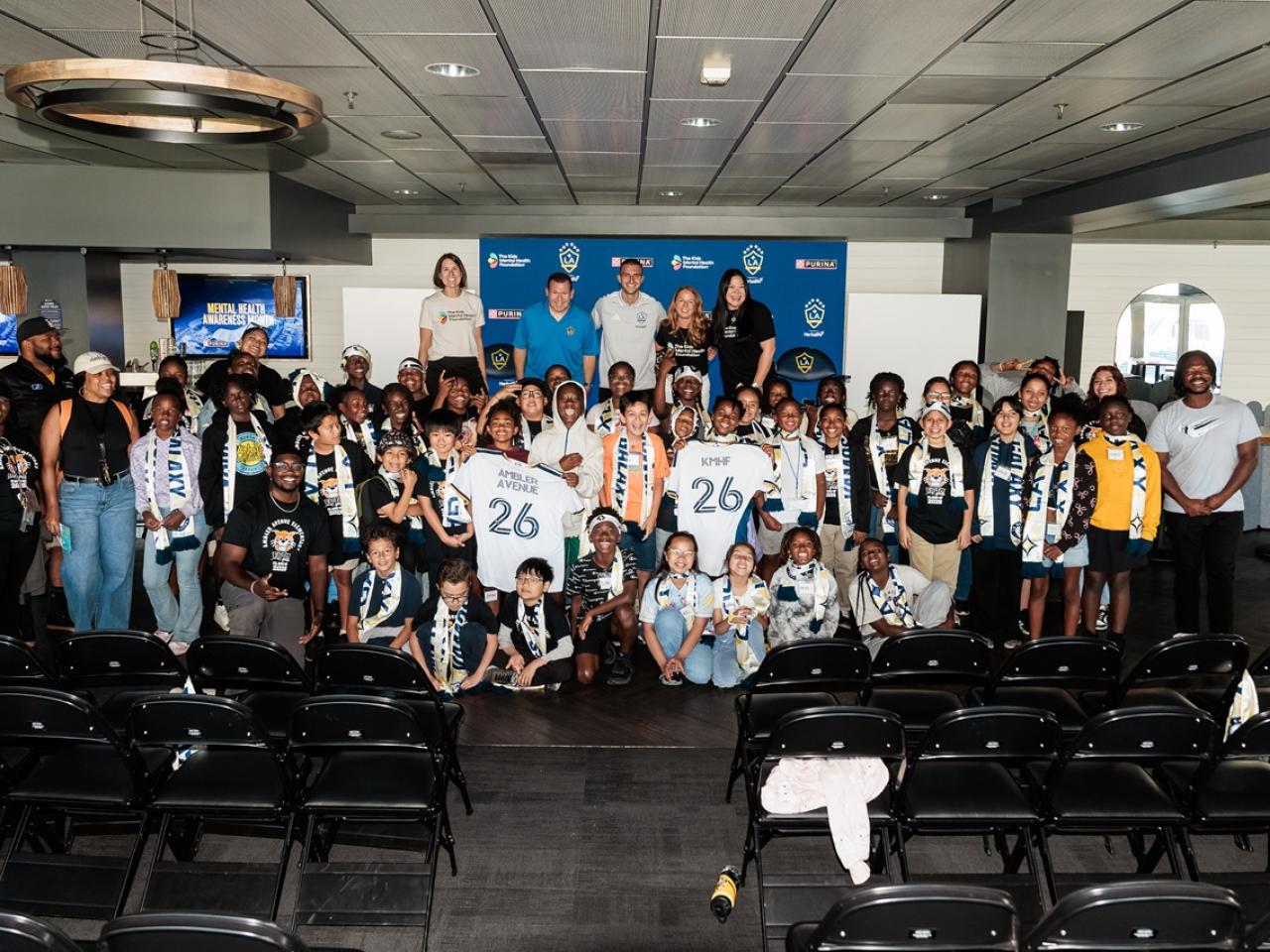 Large group of youth participants and adults pose together indoors with LA Galaxy signage and jerseys during a community event.