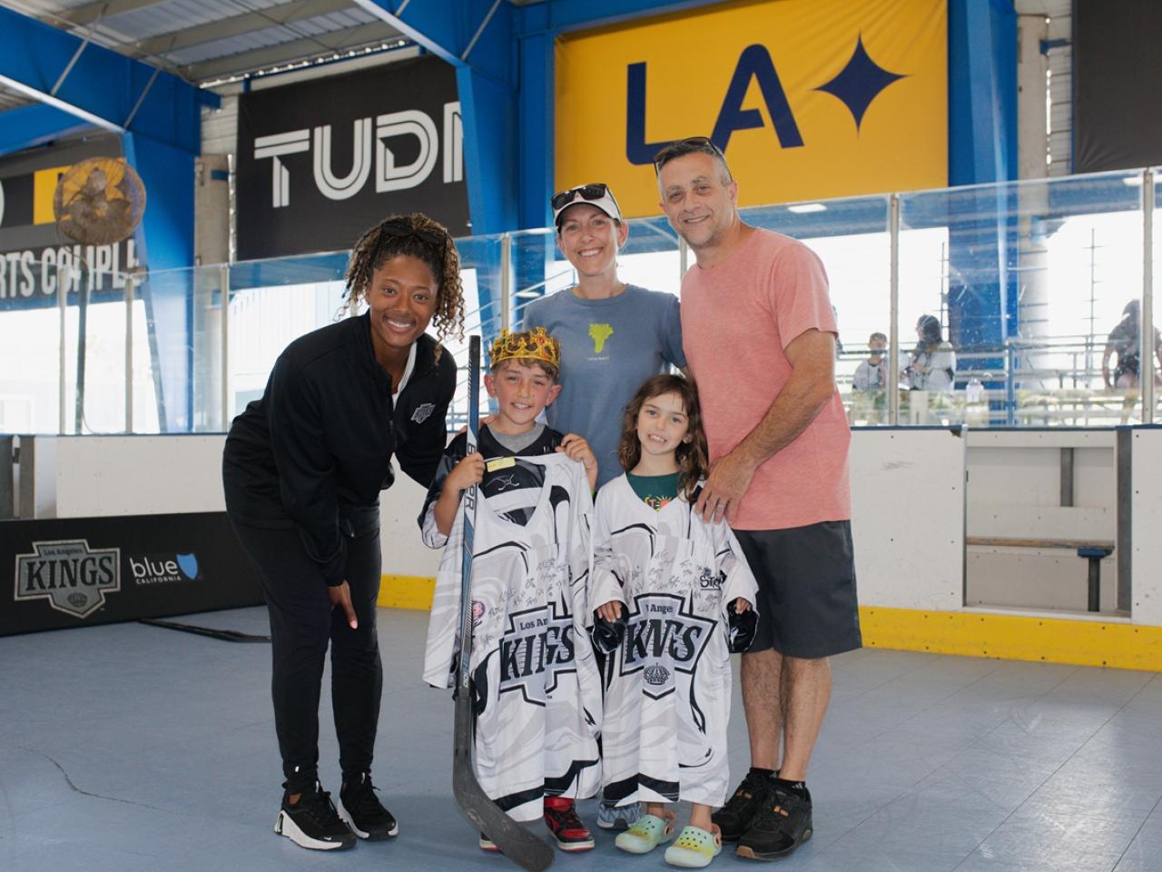 Two youth participants stand with signed LA Kings jerseys alongside family members and LA Kings Blake Bolden during a ball hockey event at an indoor sports facility.