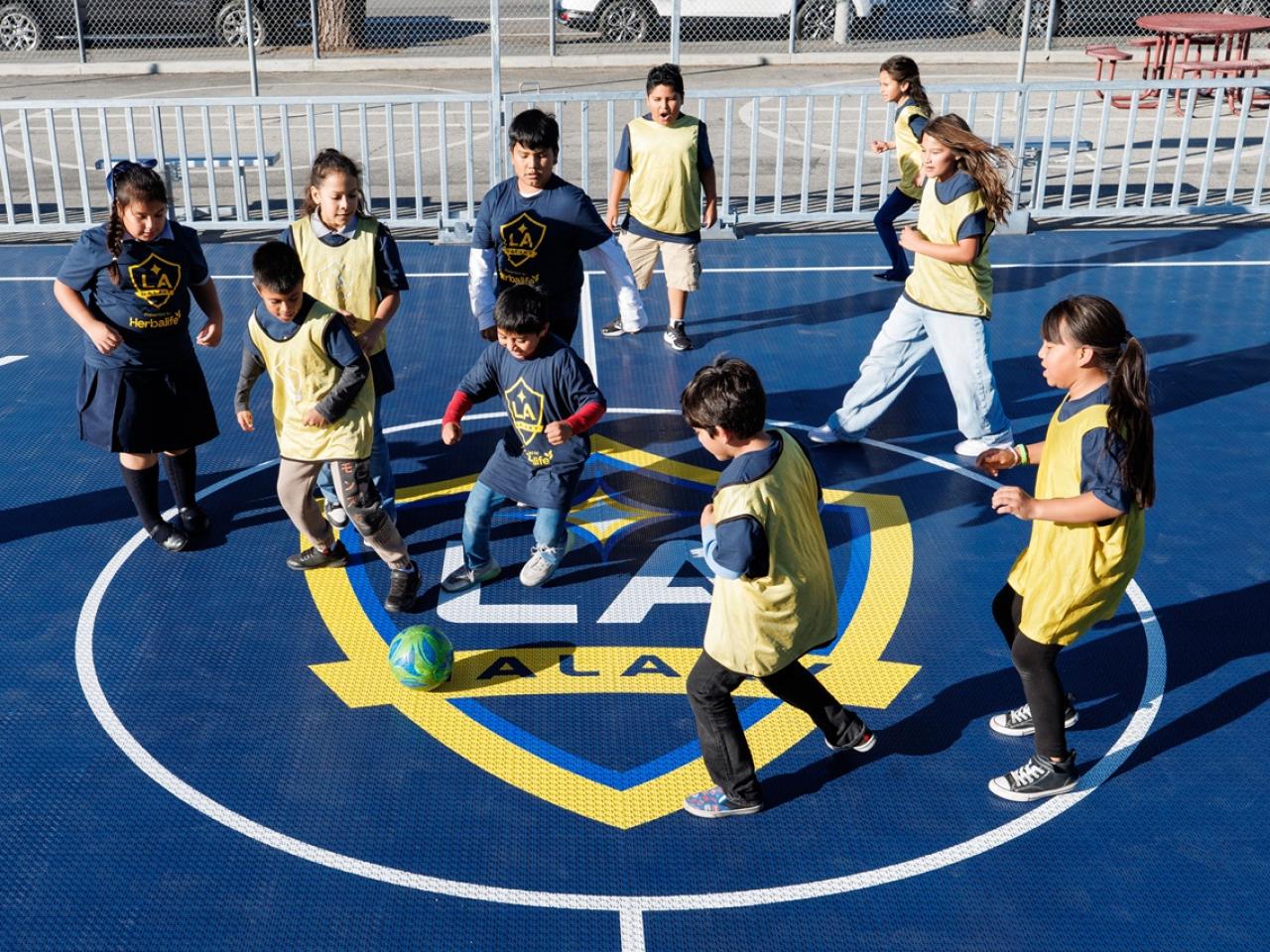 Children wearing LA Galaxy jerseys and pinnies play soccer around a ball on a branded mini-pitch court.