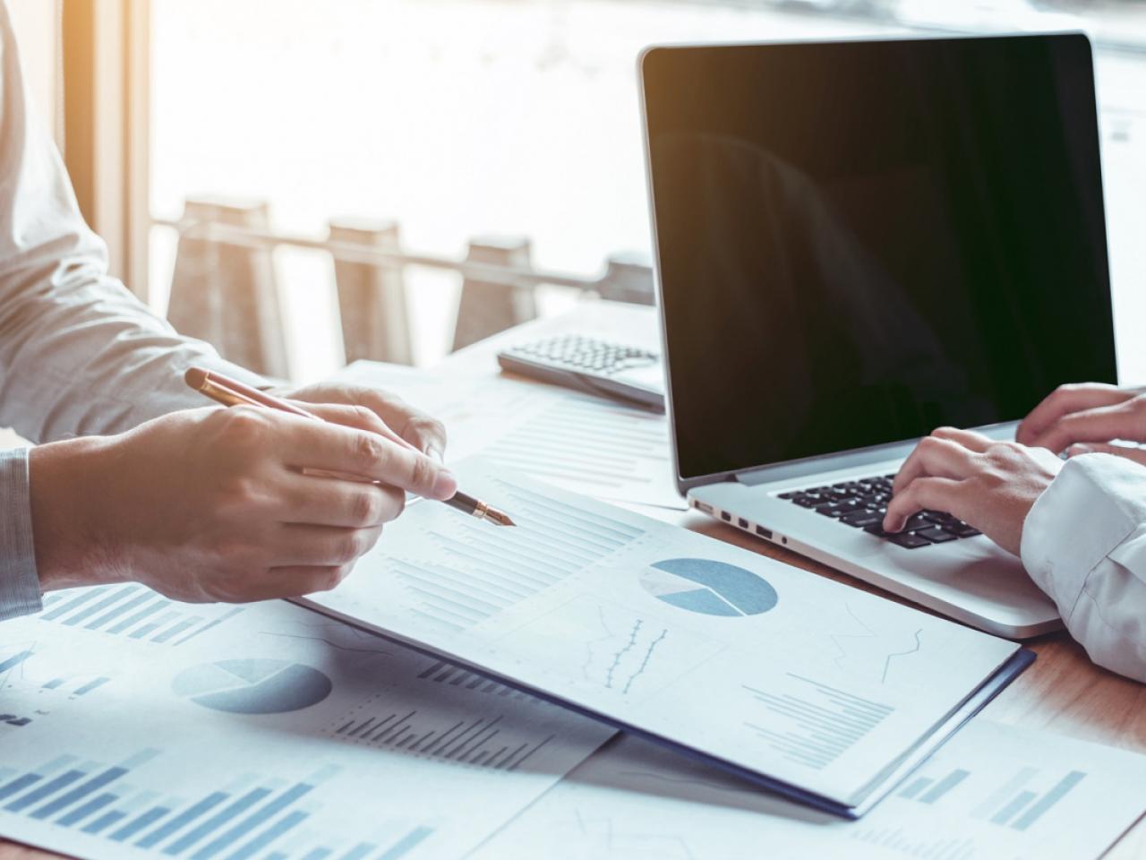 Close shot of two people working at a desk, looking at a laptop and papers