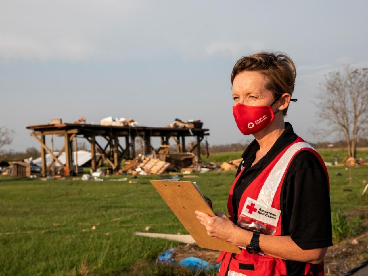 Red Cross volunteer carries a clip board in a field with debris scattered