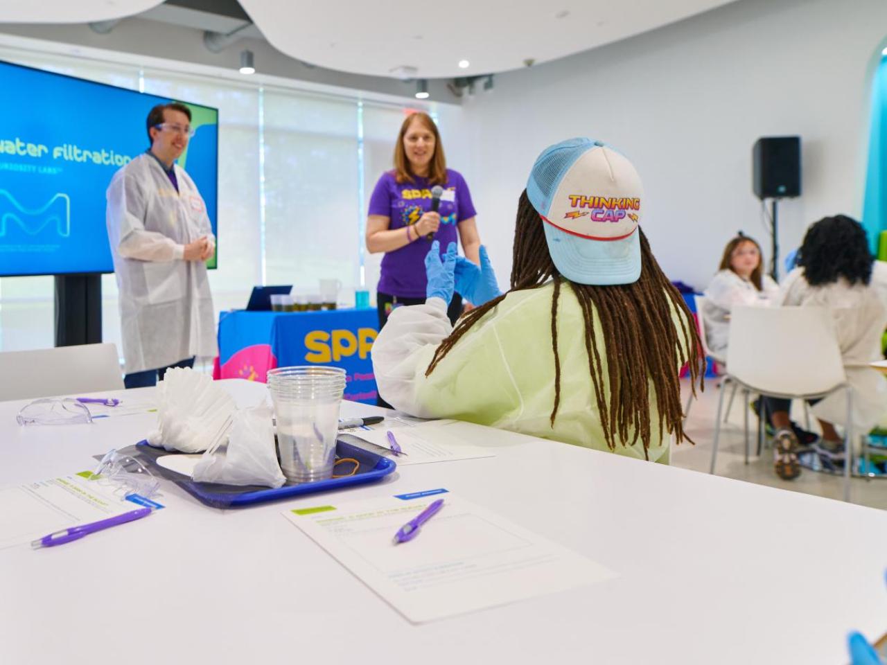 Students sit at a big table with paper and pencils while they talk with scientists at the head of a classroom.