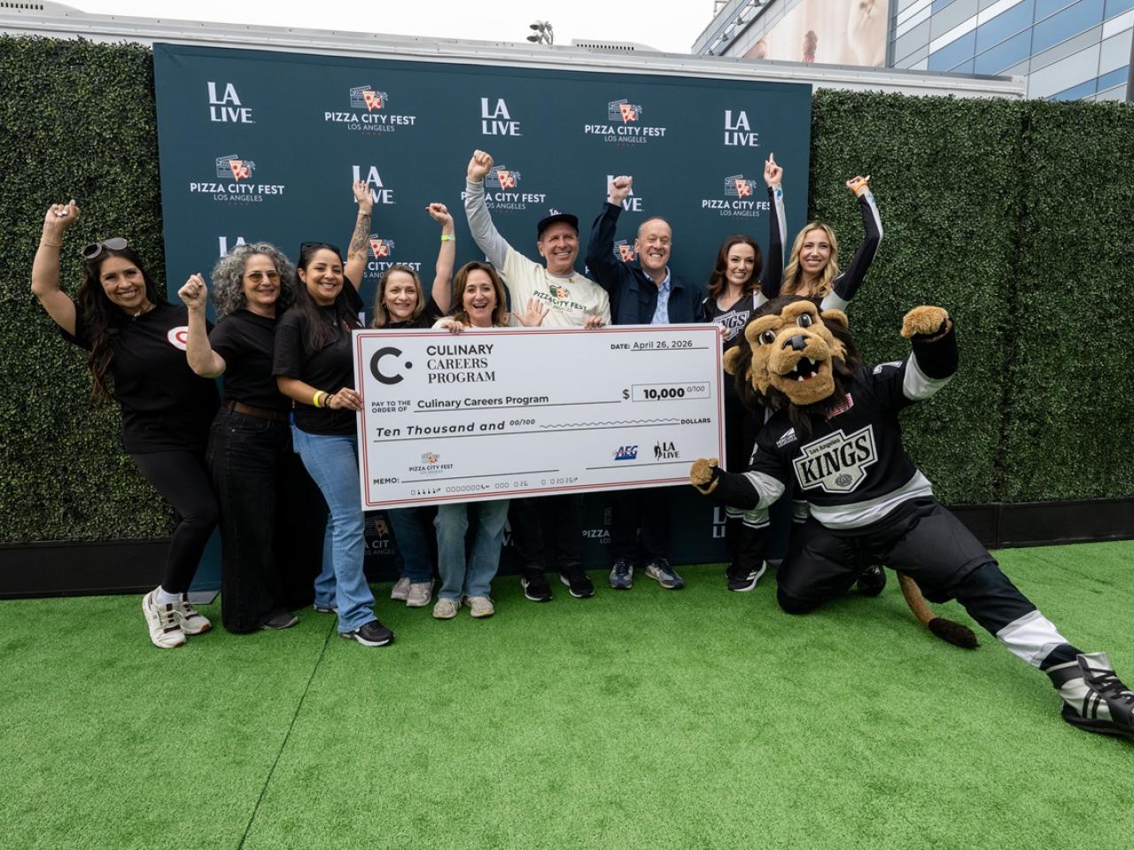 Group stands on a branded step-and-repeat holding a large donation check for the Culinary Careers Program at LA LIVE, with a mascot posing nearby.