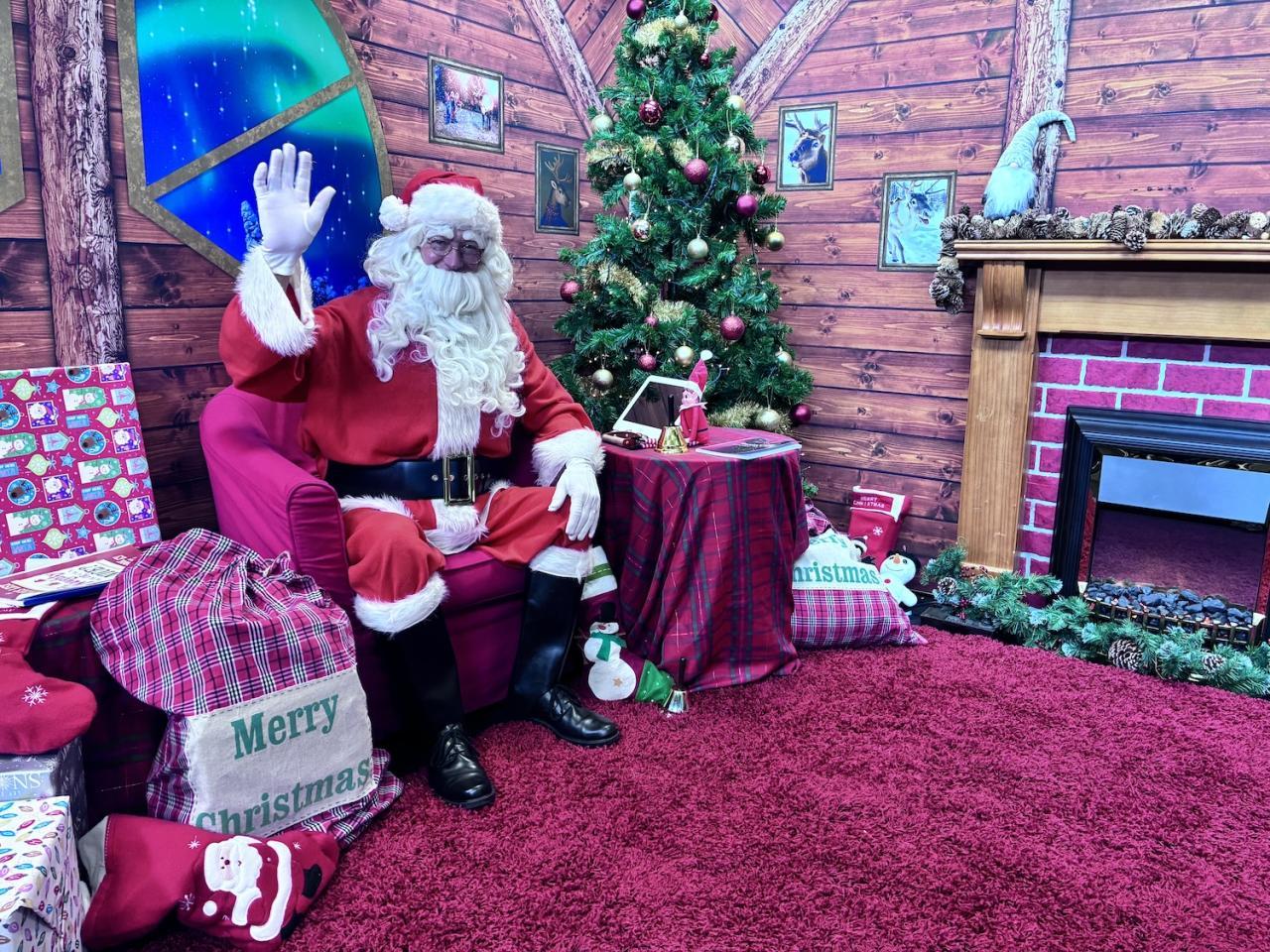 Santa waving at camera in front of Christmas tree and tablet