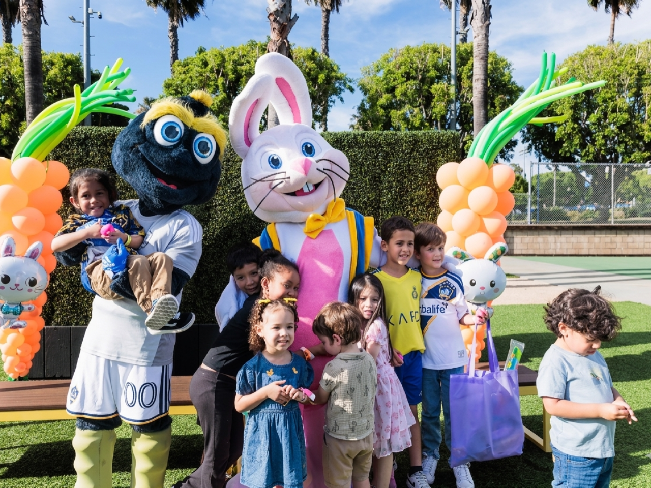Children gather with two mascots on a turf field, holding gift bags and standing beneath balloon decorations.