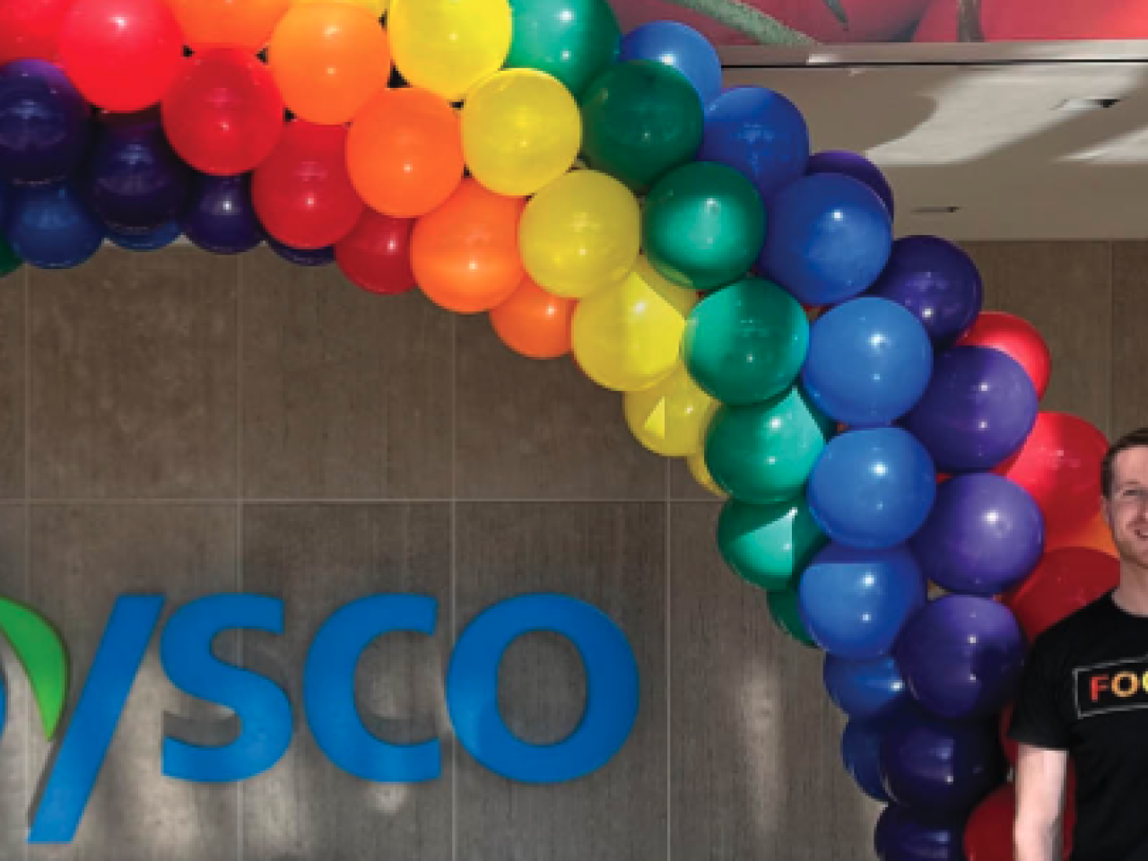 Person standing next to Sysco logo and rainbow of balloons. 