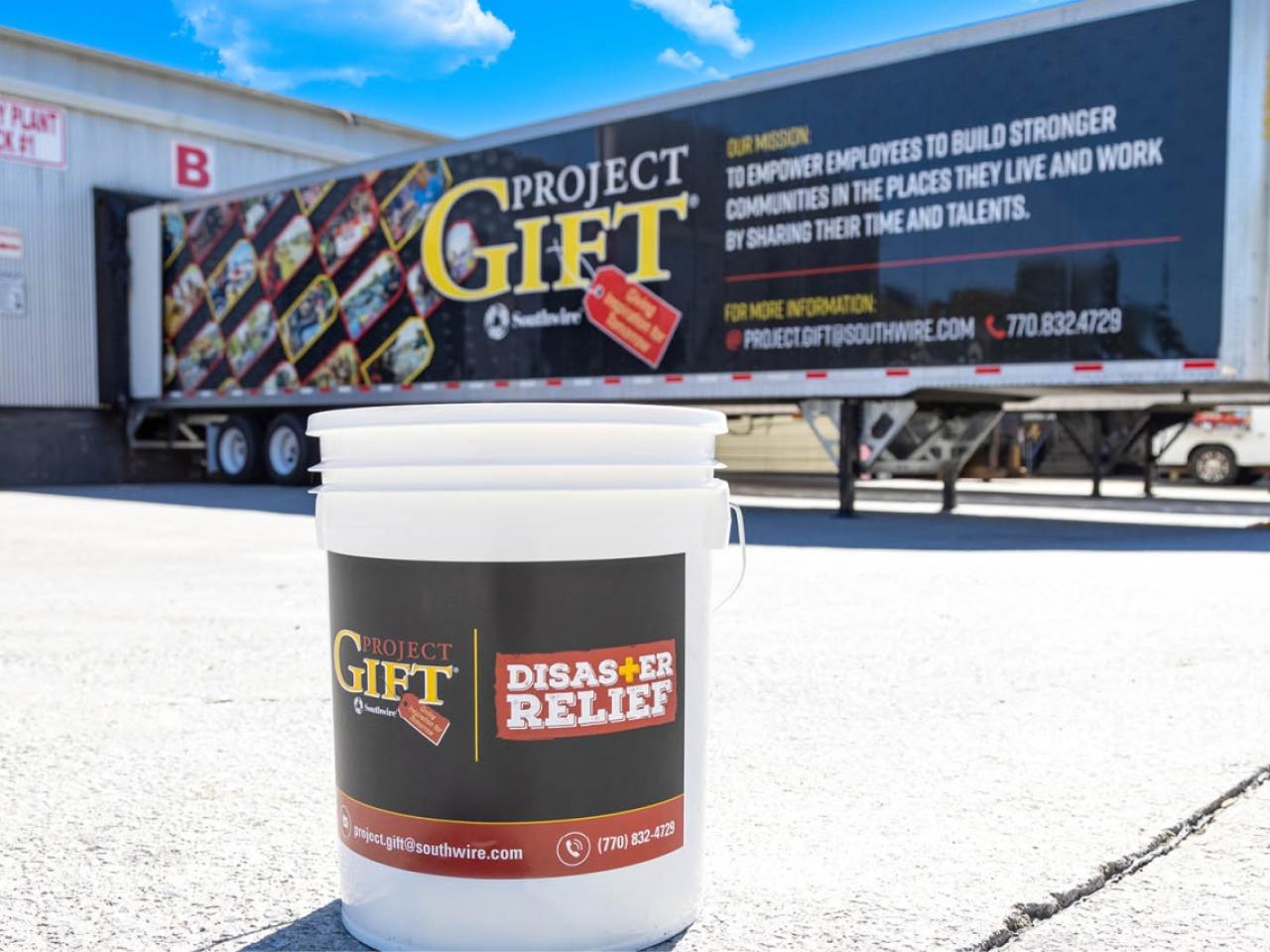 a white bucket with "GIFT disaster relief" label. A trailer in the background with "Project GIFT" on the side