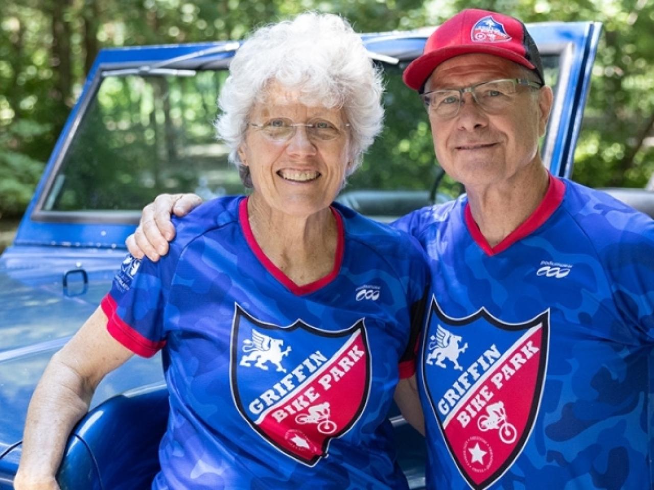 Gene and Donna Griffin, standing in front of a jeep, both wearing matching blue Griffin Bike Trail bike shirts