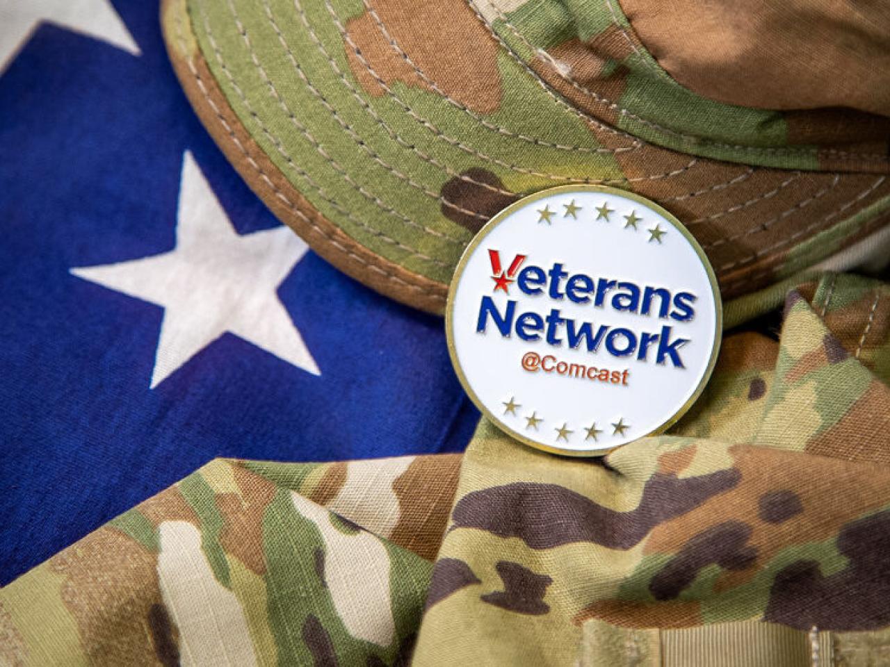 An american flag behind a camo patterned hat and shirt. A badge with "Veterans Network Comcast" on top.