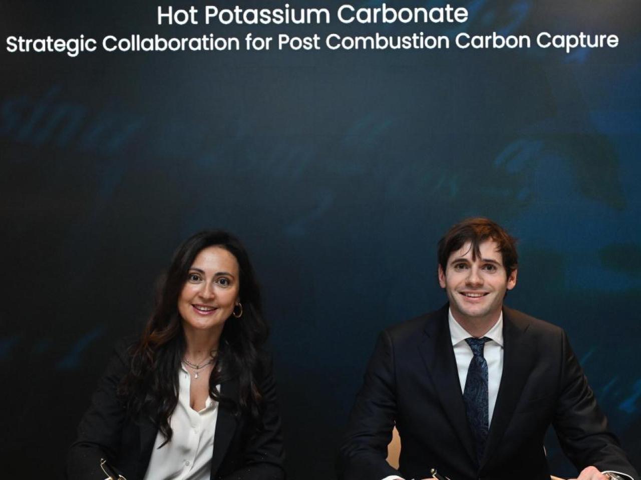 Two smiling business people sitting under the words, "Hot Potassium Carbonate Strategic Collaboration for Post Combustion Carbon Capture"