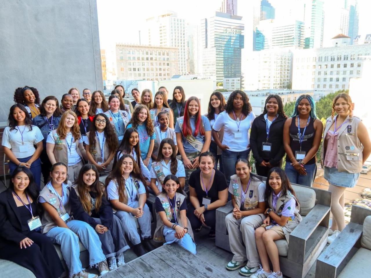 A large group of participants gathers on an outdoor terrace with city buildings in the background, posing together during a group activity.