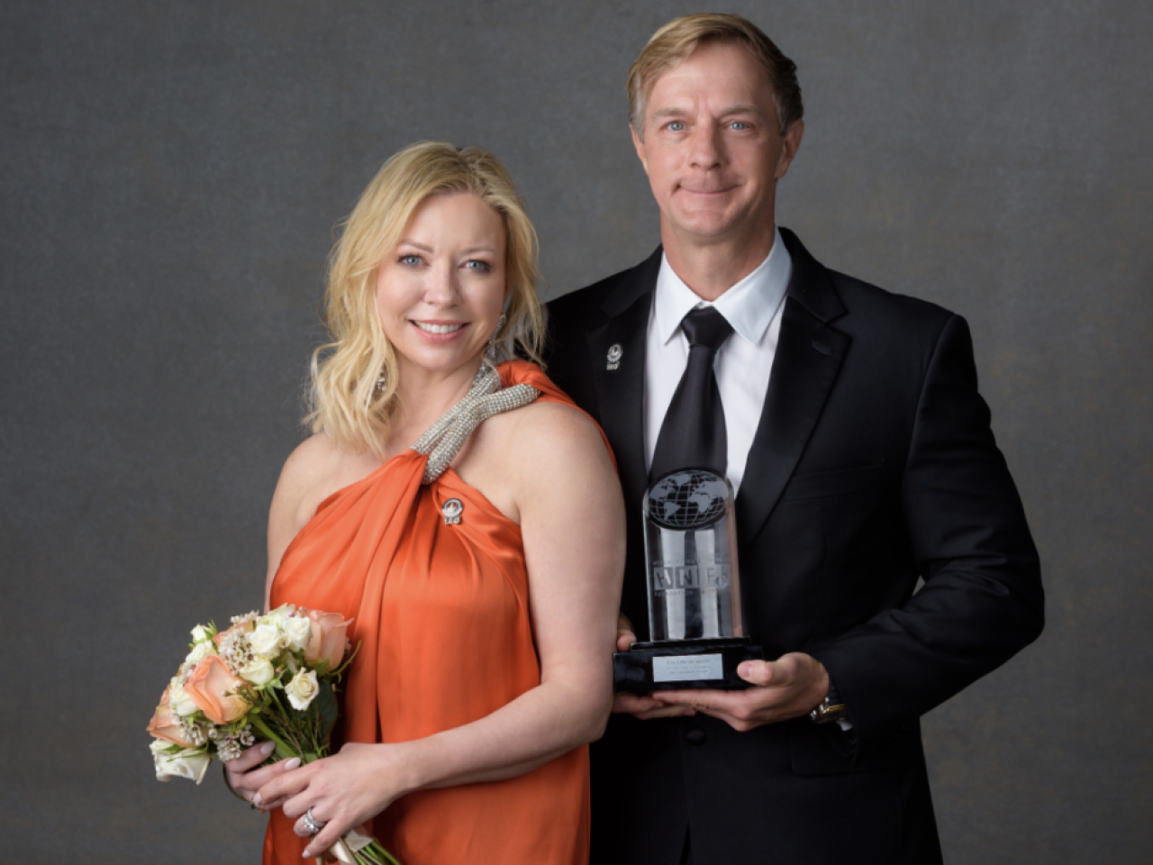 Two people stood next to each other smiling, one person holding flowers and the other holding an award