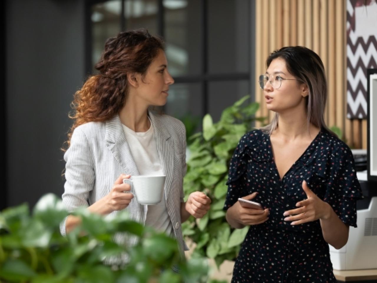 Two people talking, one with a white cup in their hand, the other has a phone. In an office setting with house plants in front and behind.
