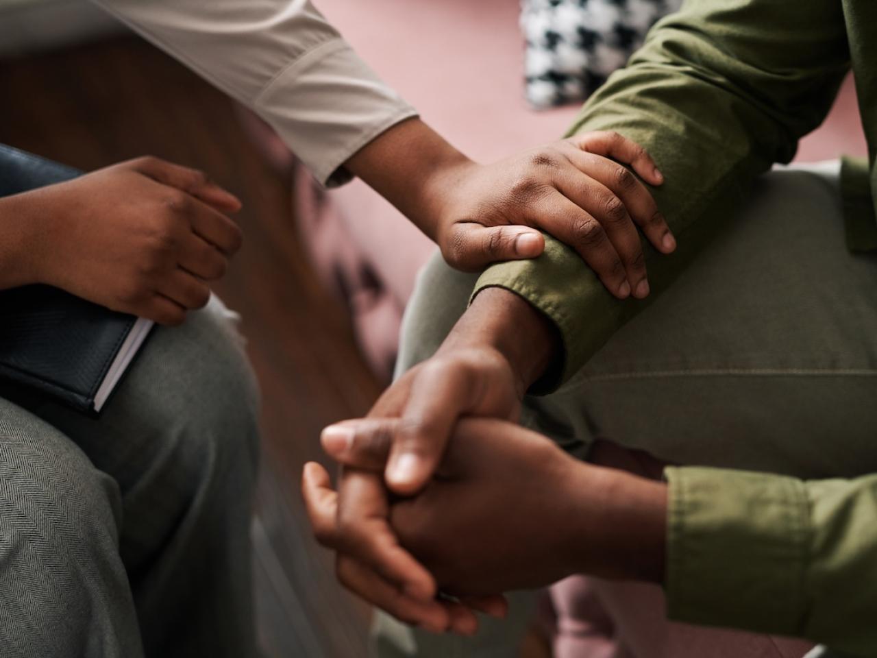 Two people seated. One with folded hands and the other placing his arm in a reassuring manner on the others arm.