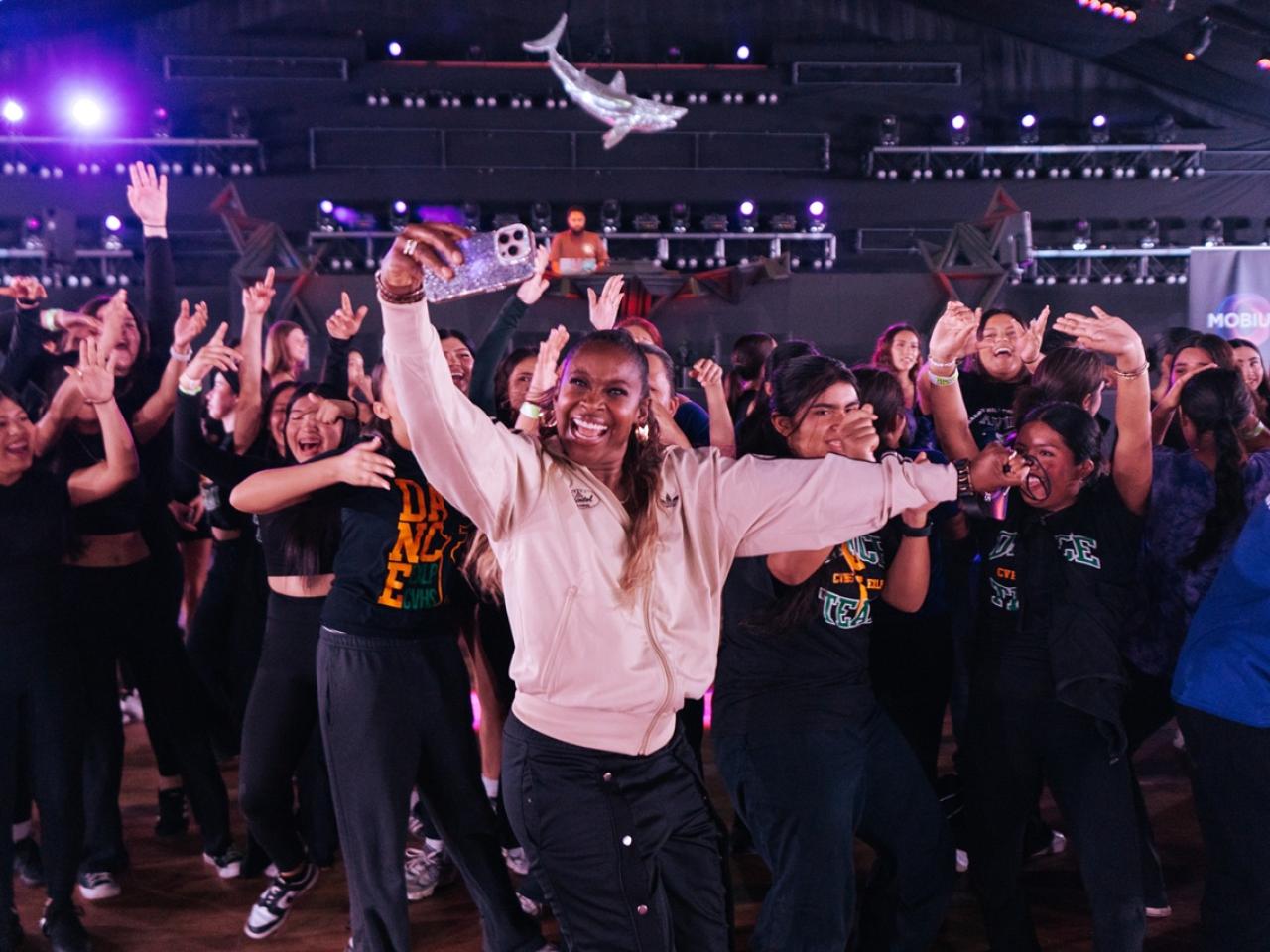 A person in a light jacket holds up a phone for a selfie while surrounded by a large group dancing with raised arms inside a concert-style venue with stage lighting overhead.