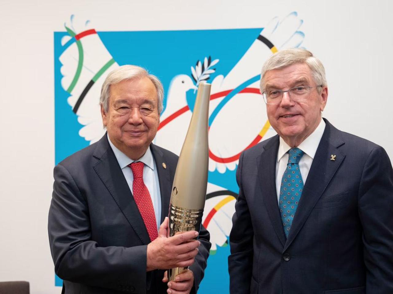 Two people stood next to each other wearing suits, the person on the left is holding the Olympic torch 