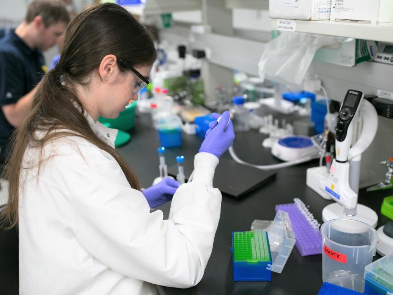 A person in protective lab gear working with pipettes and small containers at a table.