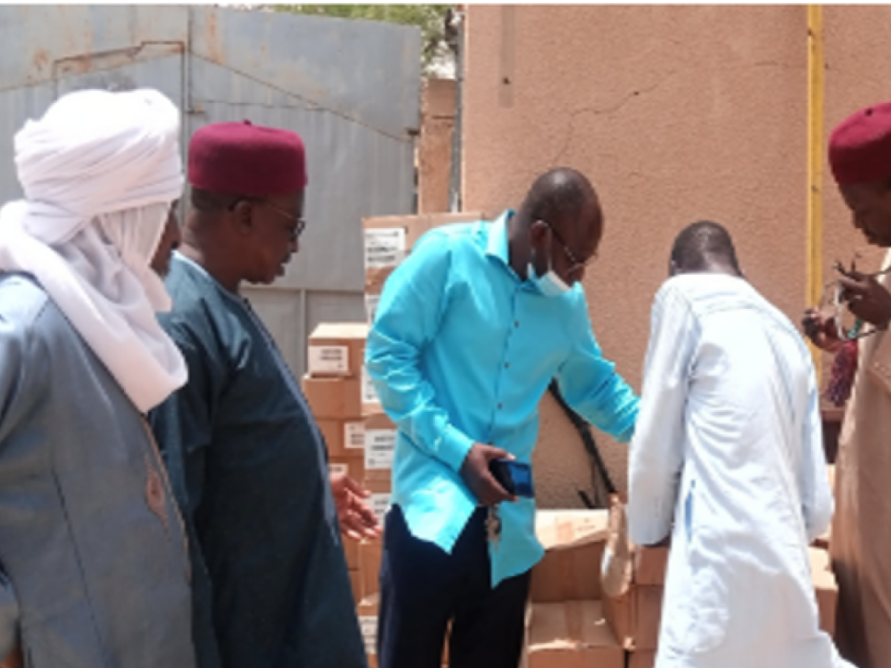Five people look over a stack of boxes outside a gated concrete wall.