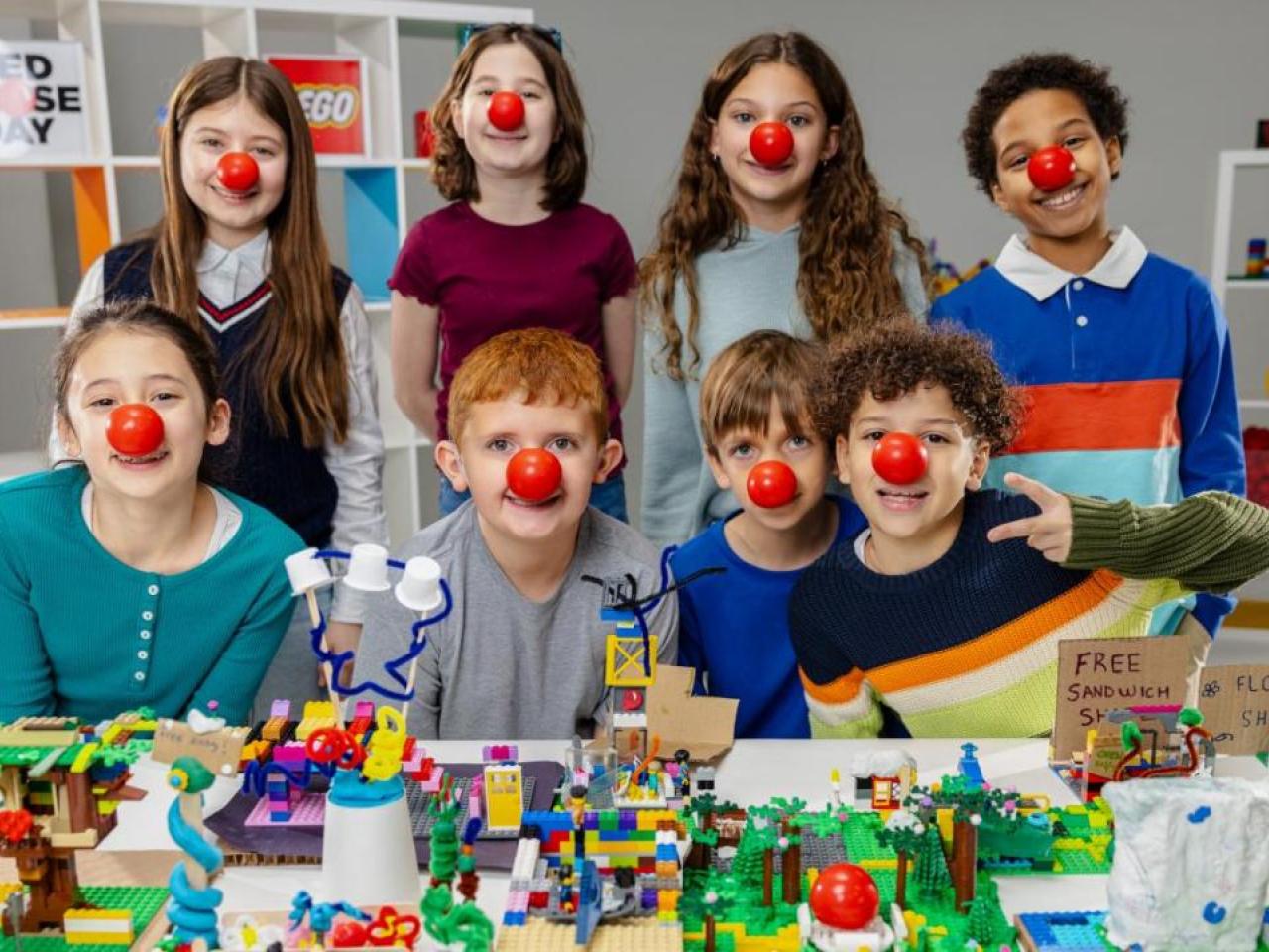 Children wearing red noses behind a table with lego models on 