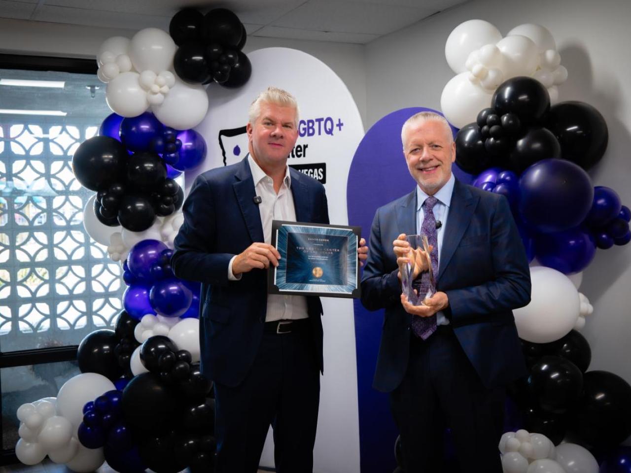 Two men standing in front of balloons. One is holding a framed certificate and one is holding an award.