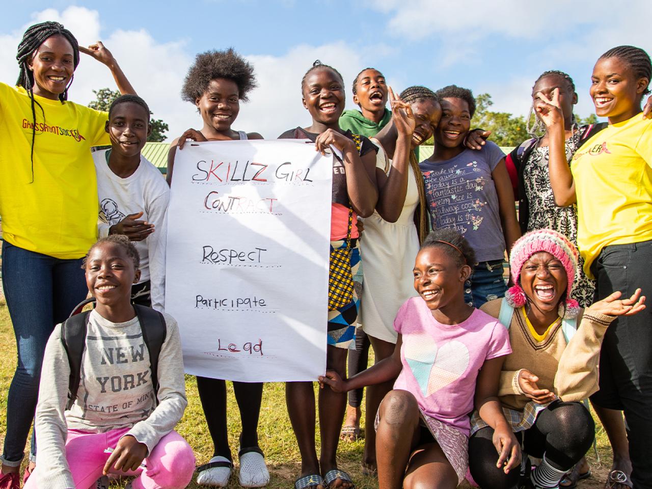 A group of youths smiling, posing for the camera on an open field, holding a sign "Skills Girl contract."